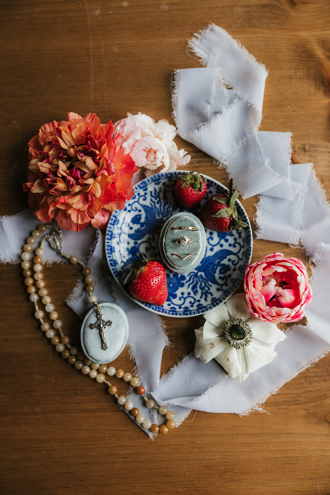 A blue and white decorative plate with strawberries and a porcelain egg, surrounded by flowers, beads, and white fabric on a wooden surface for spring wedding inspo