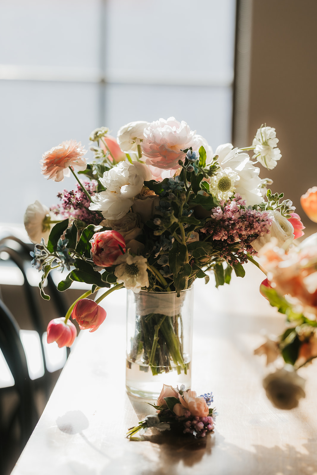A glass vase filled with assorted fresh flowers, including pink, white, and purple blooms, sits on a sunlit table.