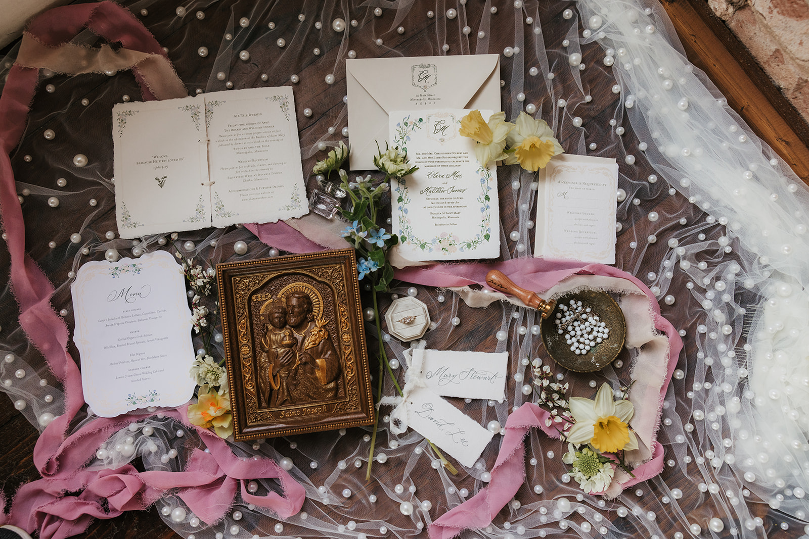 Wedding invitations, stationery, and a religious icon are arranged on a dotted sheer fabric with flowers and ribbons surrounding the items.