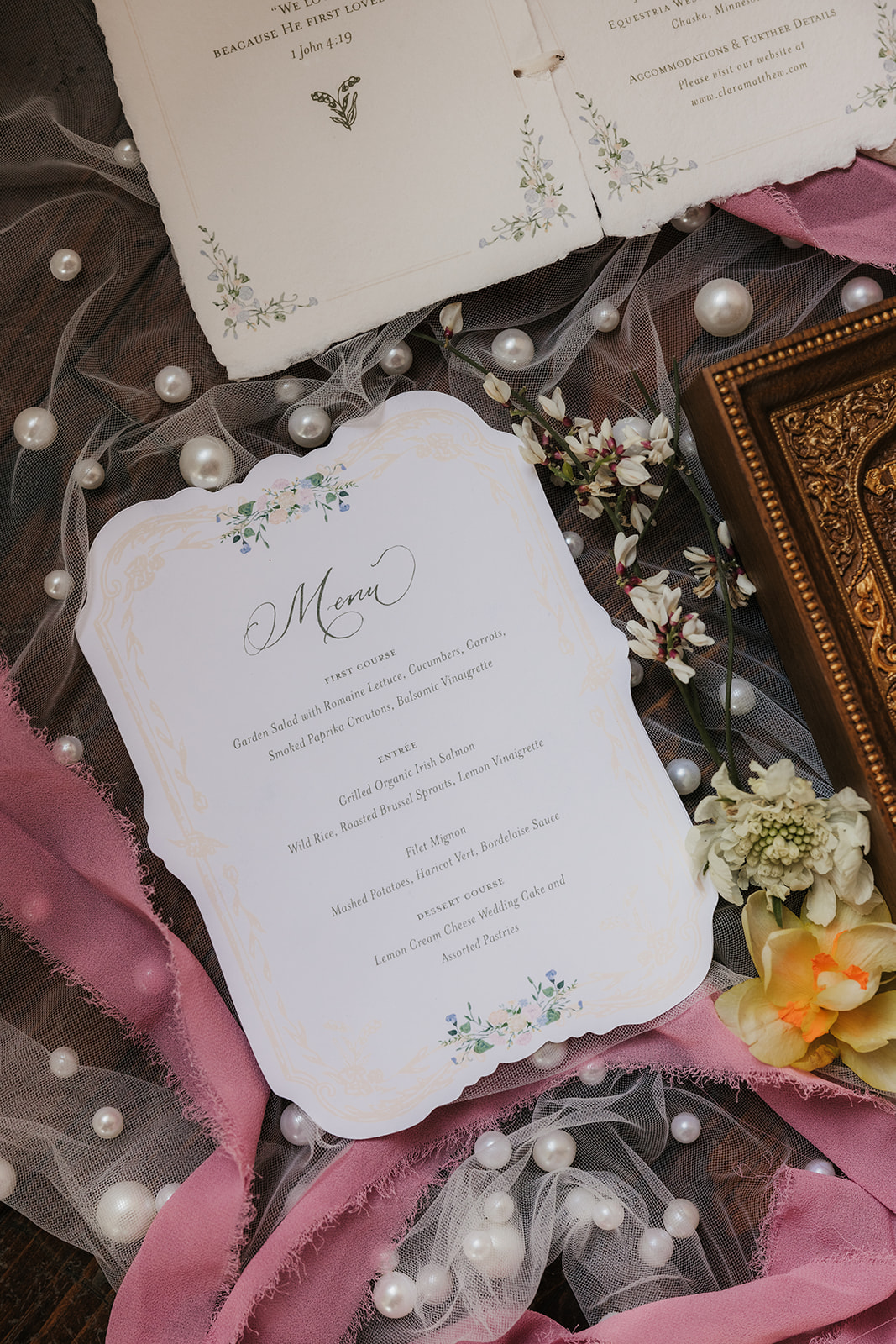 Wedding invitations, stationery, and a religious icon are arranged on a dotted sheer fabric with flowers and ribbons surrounding the items.