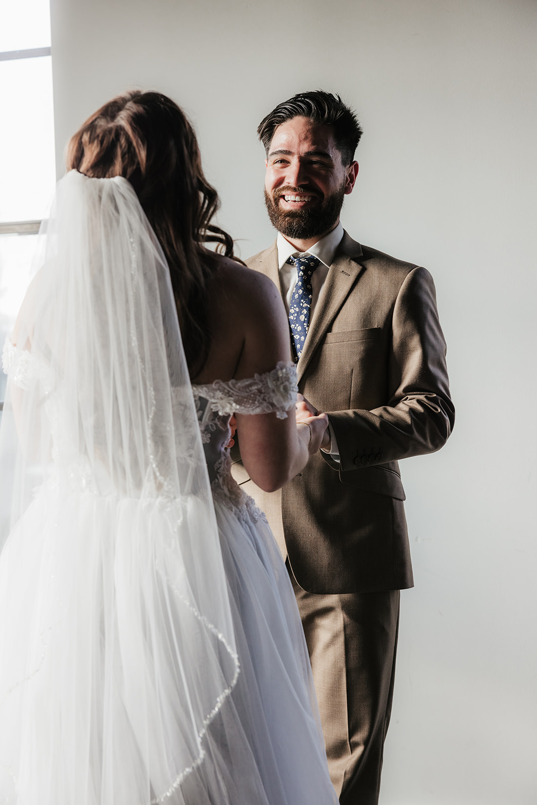 A bride in a white gown and veil stands with a groom in a tan suit in a sunlit room with large windows and a floral arrangement on a cart in the background.