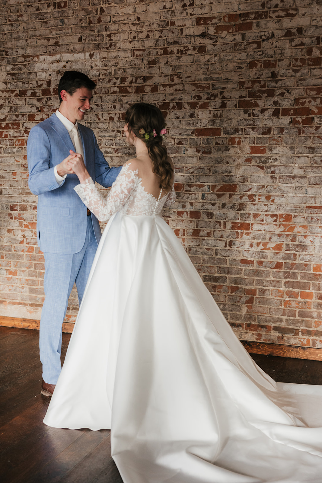 A bride in a white gown and veil stands with a groom in a tan suit in a sunlit room with large windows and a floral arrangement on a cart in the background.