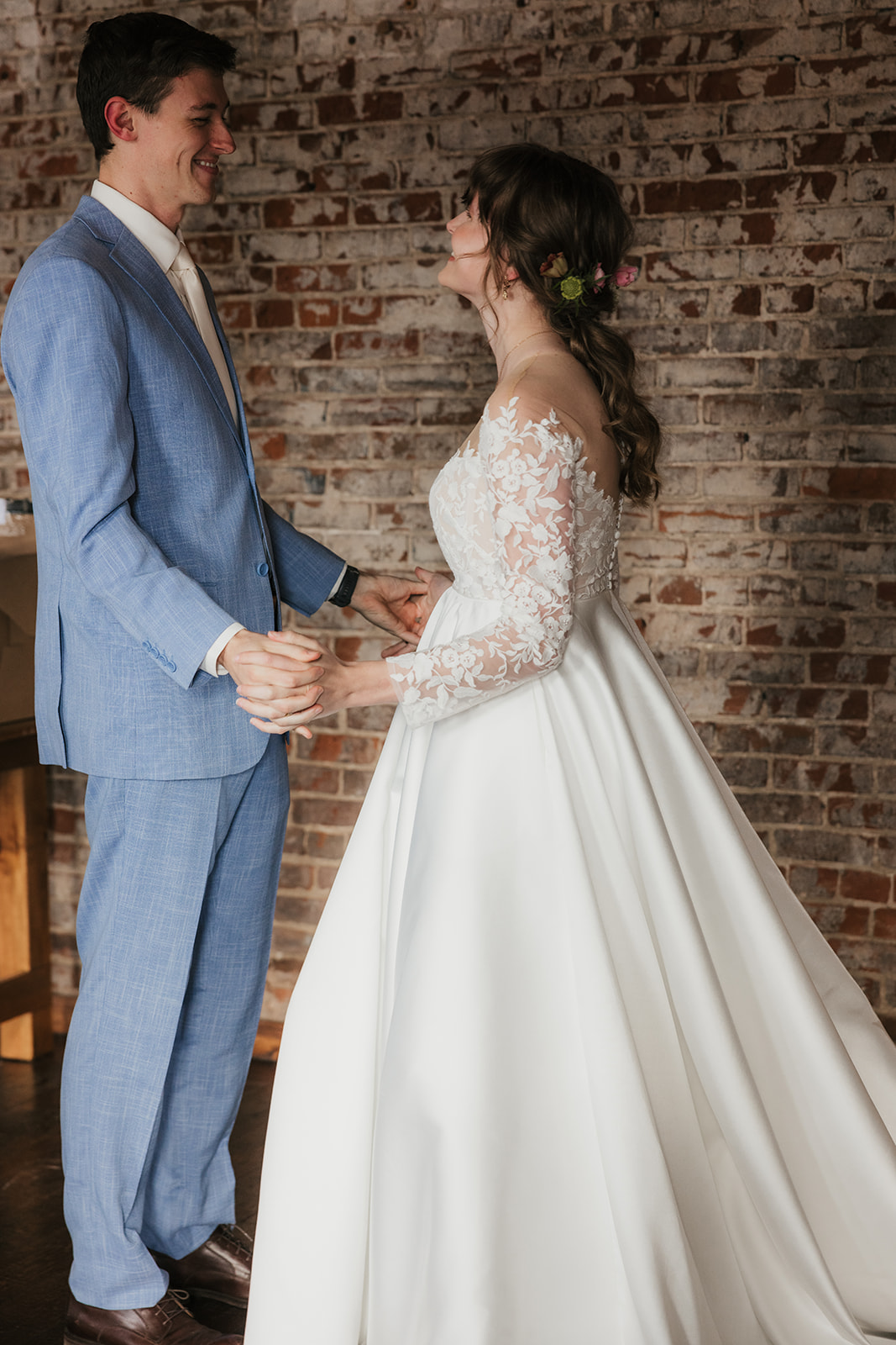 A bride in a white gown and veil stands with a groom in a tan suit in a sunlit room with large windows and a floral arrangement on a cart in the background.