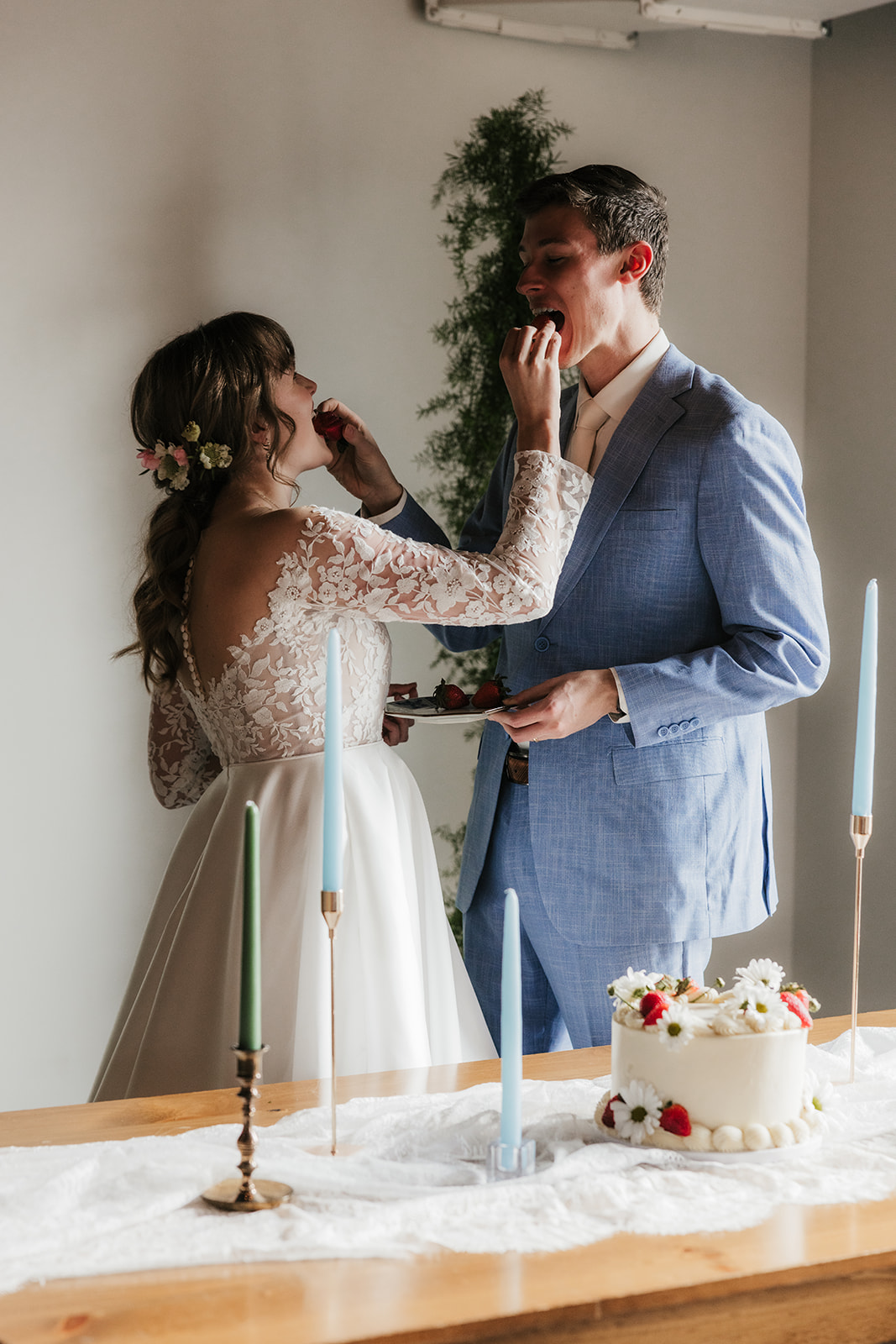Bride and groom feed each other cake at a wedding, standing near a decorated table with candles and a white cake.