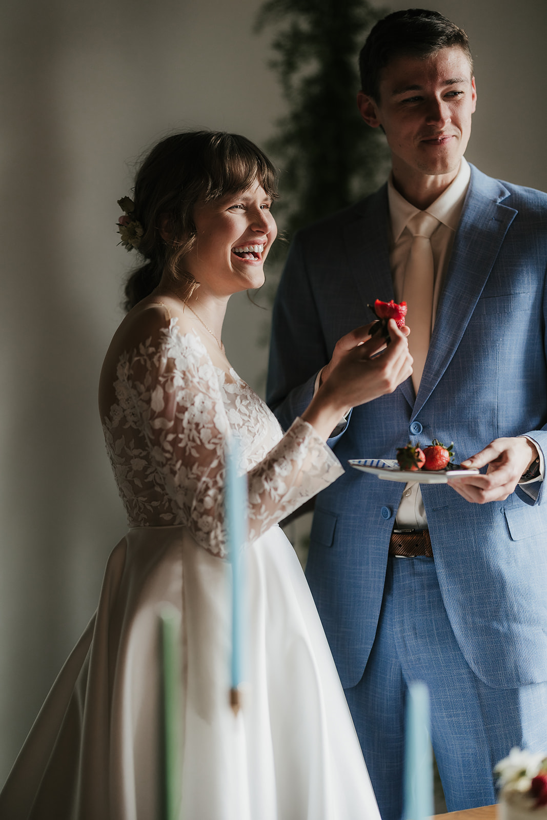 Bride and groom feed each other cake at a wedding, standing near a decorated table with candles and a white cake.