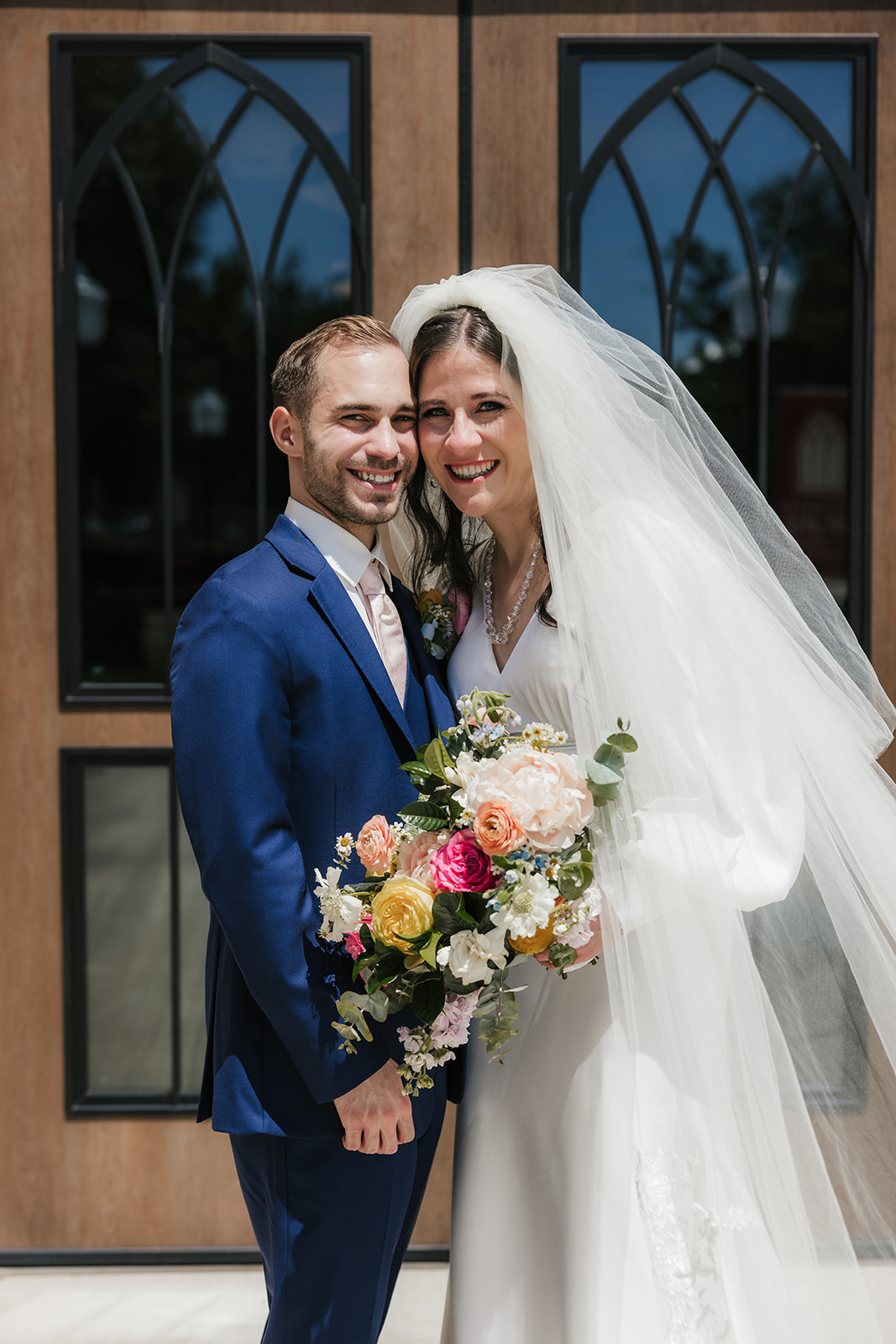 A bride in a white dress and veil stands beside a groom in a blue suit, both smiling and holding a colorful bouquet in front of wooden doors with glass panels for a Wedding at St. Thomas More University Parish