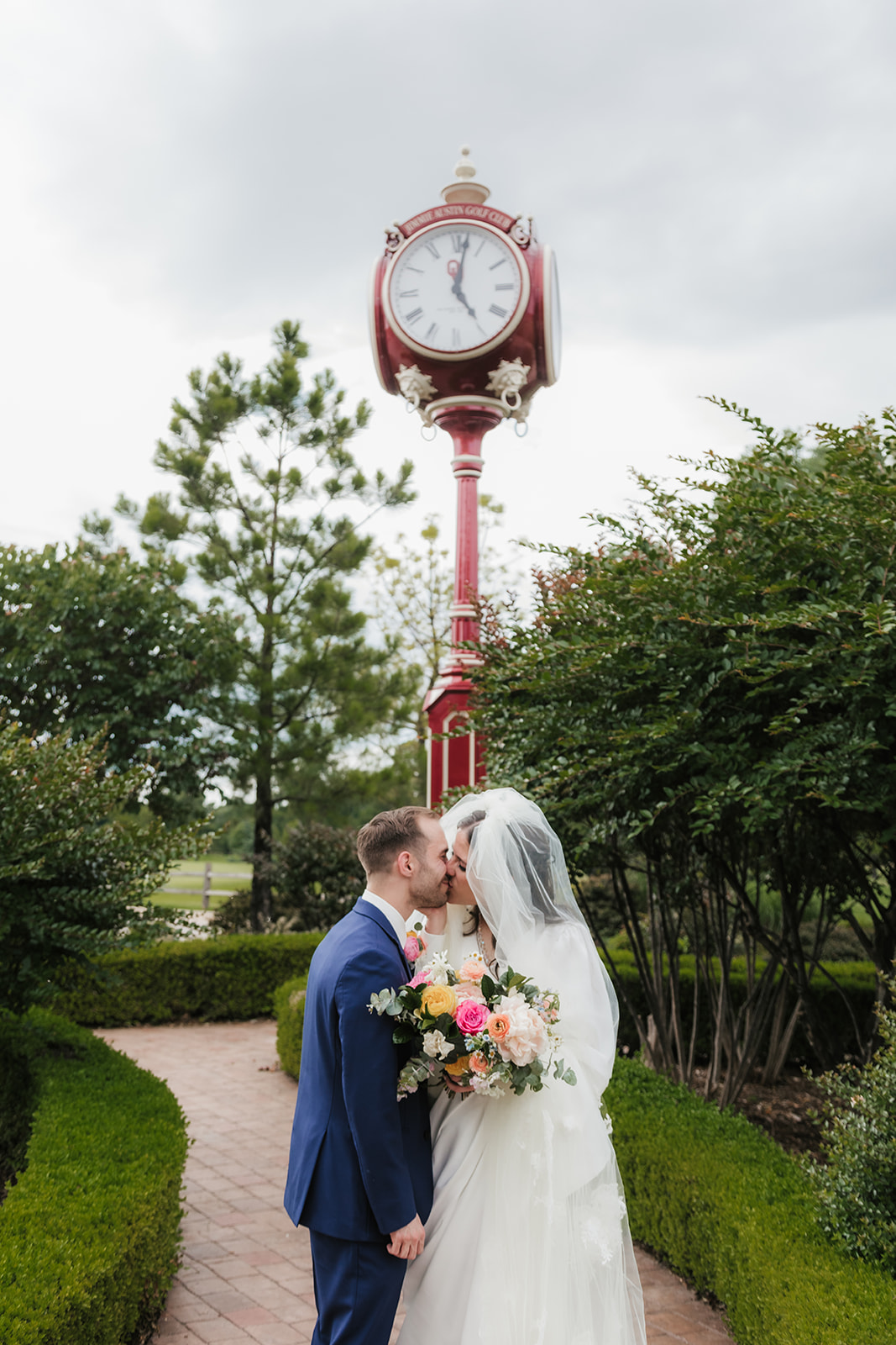 A bride in a white dress and veil stands beside a groom in a blue suit, both smiling and holding a colorful bouquet in front of wooden doors with glass panels for a Wedding at St. Thomas More University Parish