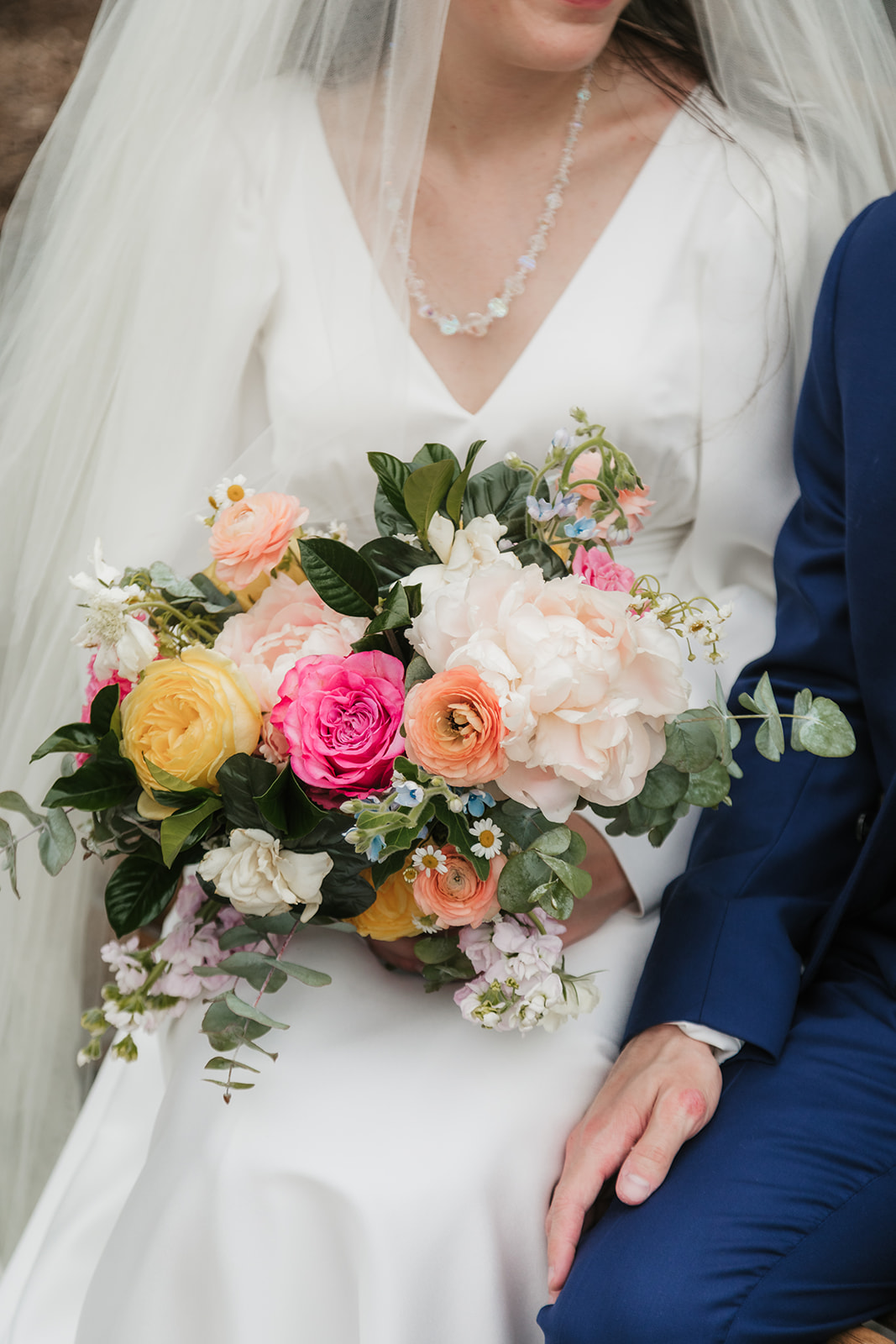 A bride in a white dress and veil stands beside a groom in a blue suit, both smiling and holding a colorful bouquet in front of wooden doors with glass panels for a Wedding at St. Thomas More University Parish