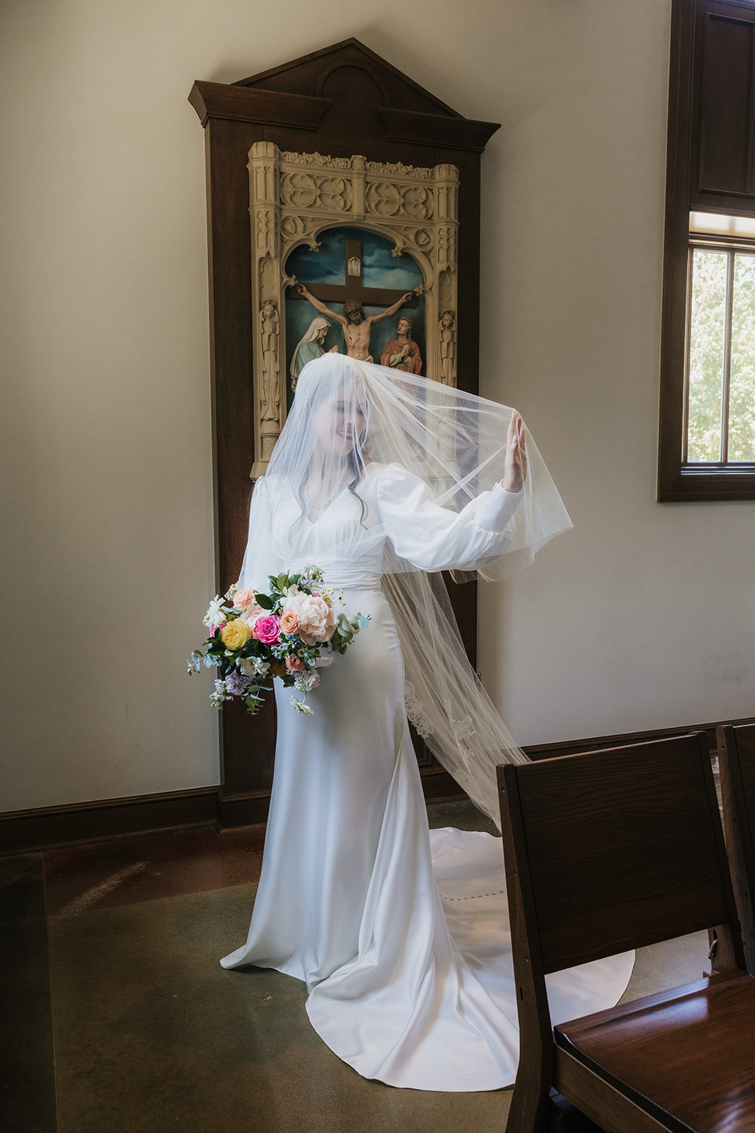 A bride in a white gown stands by a window in a church, holding a bouquet and looking outside, with sunlight streaming in for a Wedding at St. Thomas More University Parish