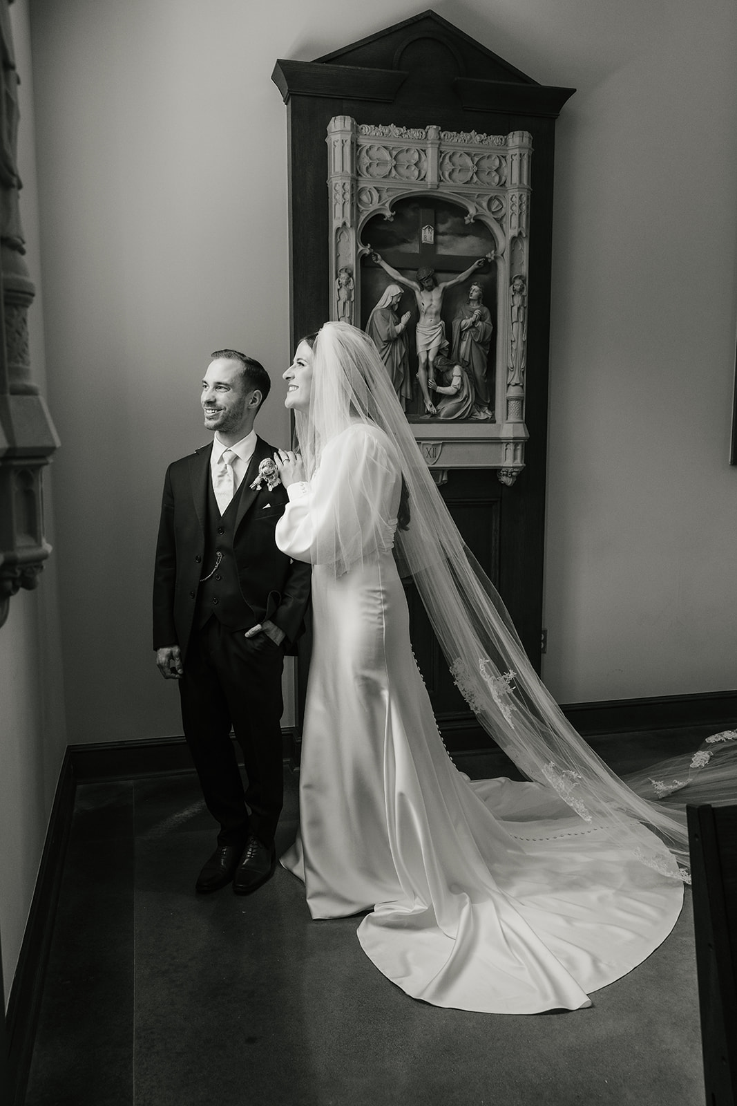 A bride in a long gown and veil stands beside a groom in a suit, both looking to the side, in an indoor setting with framed artwork on the wall for a Wedding at St. Thomas More University Parish