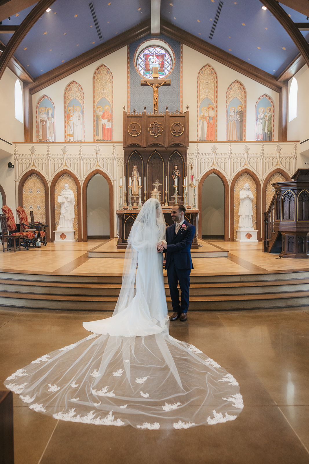 A bride and groom stand together at the altar of a church, with the bride wearing a long white dress and veil. The church interior features religious artwork and stained glass for a Wedding at St. Thomas More University Parish