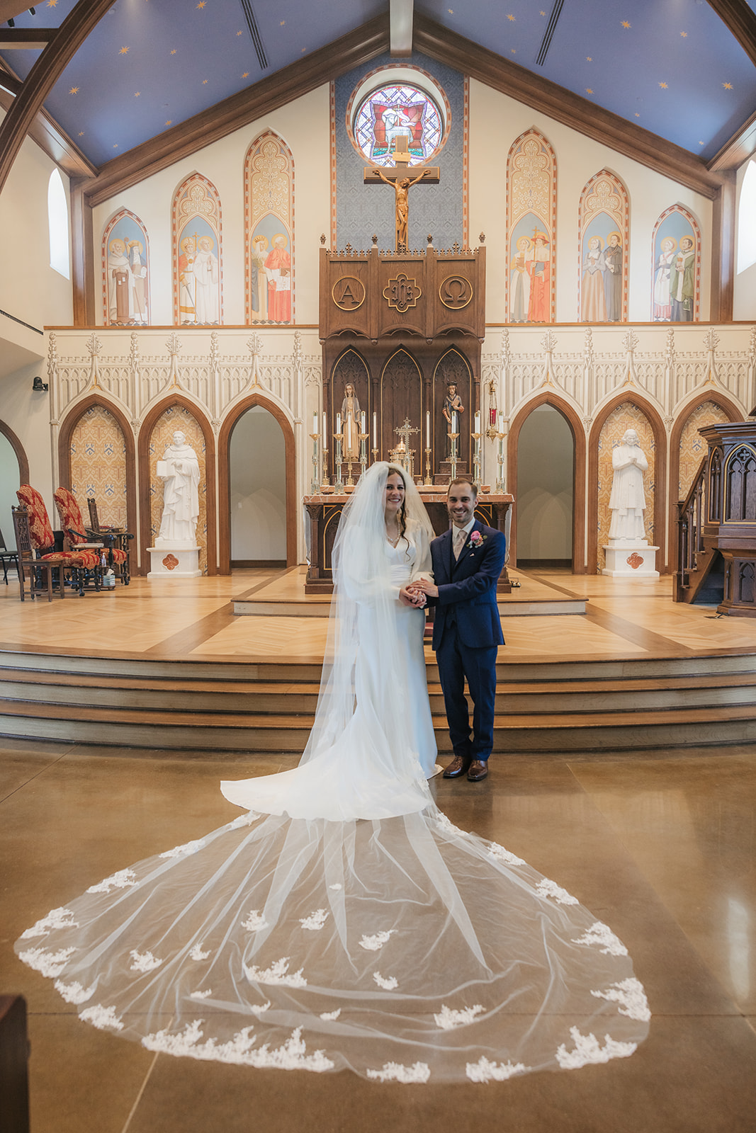 A bride and groom stand together at the altar of a church, with the bride wearing a long white dress and veil. The church interior features religious artwork and stained glass for a Wedding at St. Thomas More University Parish
