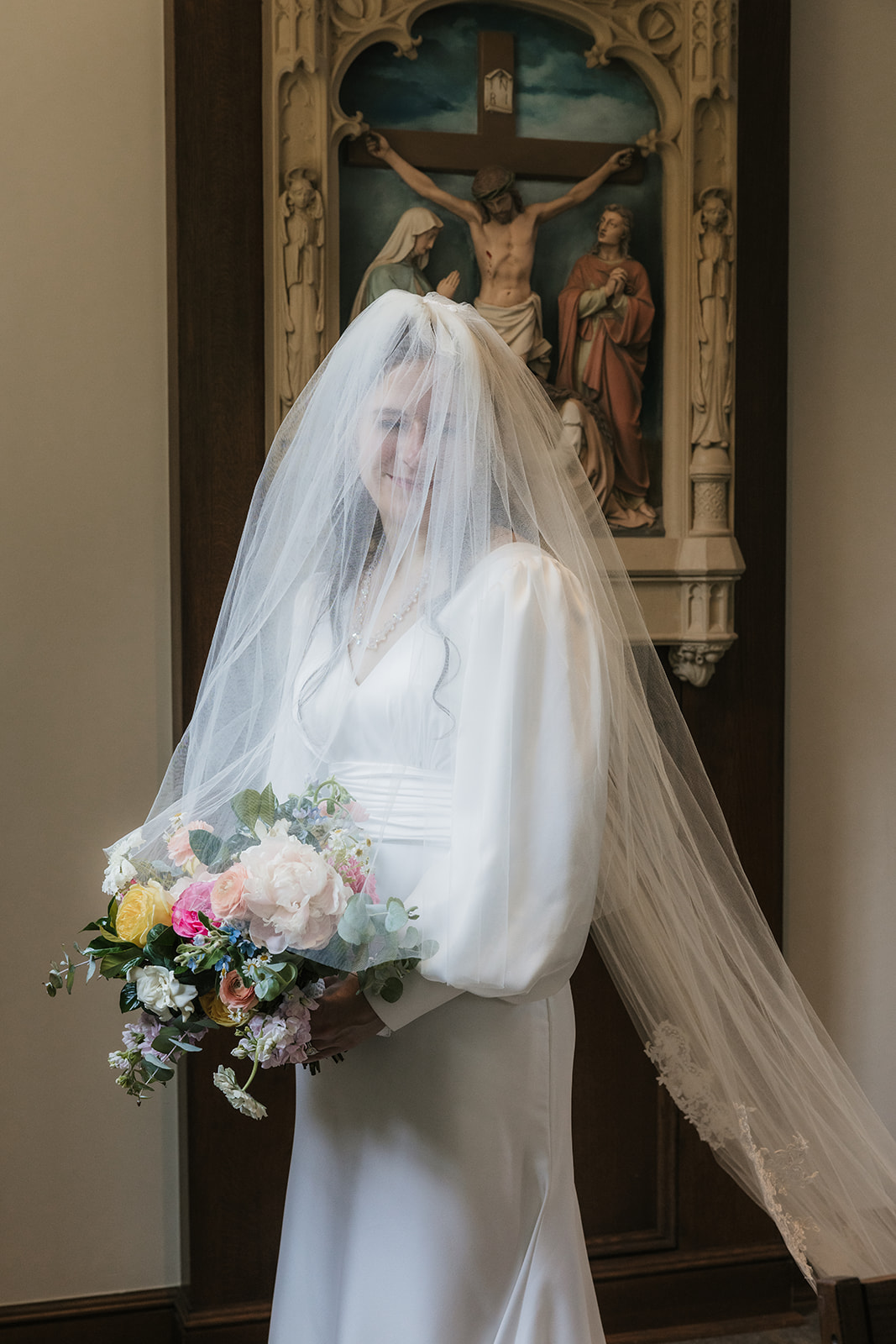 A bride in a white gown stands by a window in a church, holding a bouquet and looking outside, with sunlight streaming in for a Wedding at St. Thomas More University Parish