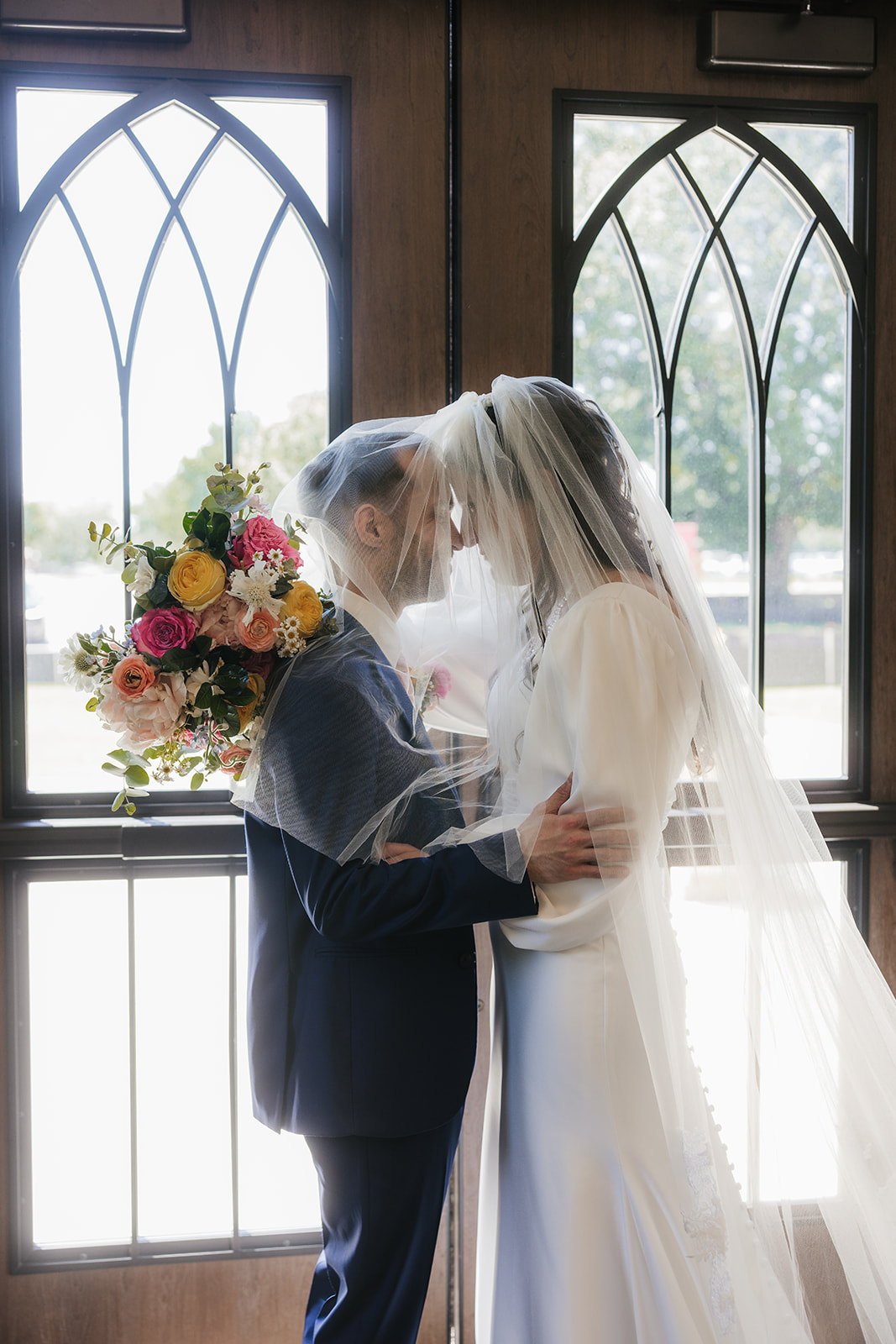 bride and groom in wedding attire stand face to face under a shared veil, embracing in front of double doors with a bouquet of flowers in the background for aWedding at St. Thomas More University Parish