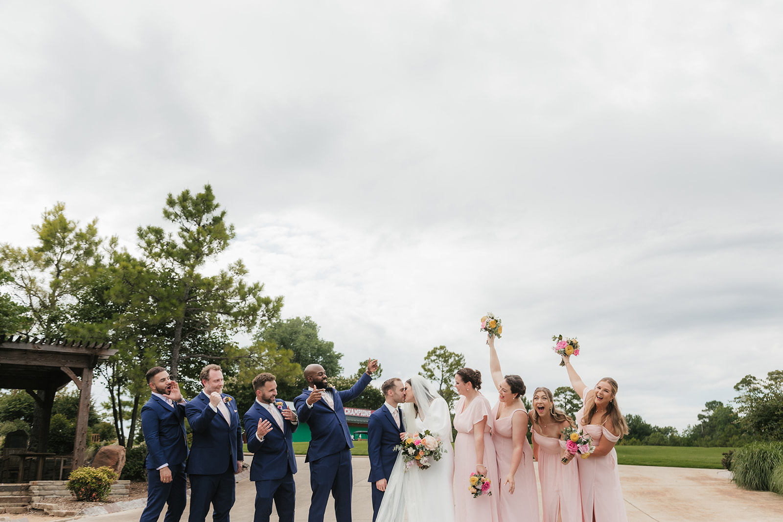 A bride and groom stand with four groomsmen in blue suits and five bridesmaids in light pink dresses, holding bouquets, outdoors on a paved area for a Wedding at St. Thomas More University Parish