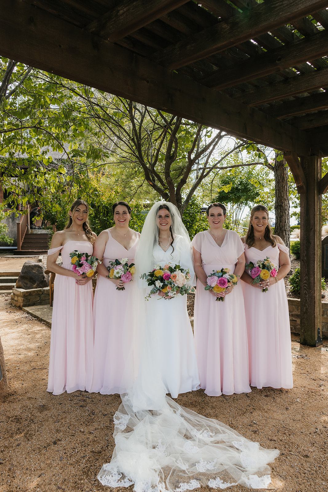 A bride and groom stand with four groomsmen in blue suits and five bridesmaids in light pink dresses, holding bouquets, outdoors on a paved area for a Wedding at St. Thomas More University Parish