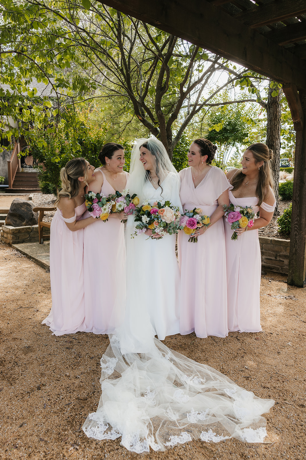 A bride and groom stand with four groomsmen in blue suits and five bridesmaids in light pink dresses, holding bouquets, outdoors on a paved area for a Wedding at St. Thomas More University Parish