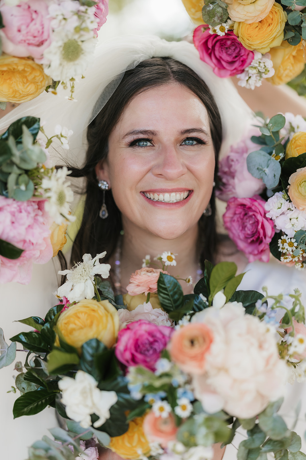 Smiling bride holding a bouquet of colorful flowers, surrounded by additional floral arrangements.