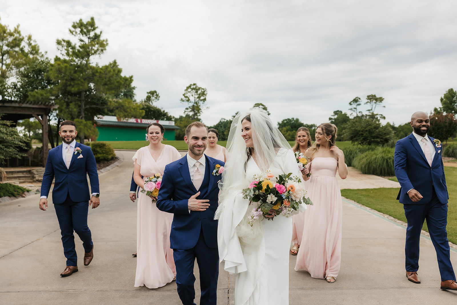 A bride and groom walk outdoors with bridesmaids in pink dresses and groomsmen in blue suits on a cloudy day for aWedding at St. Thomas More University Parish