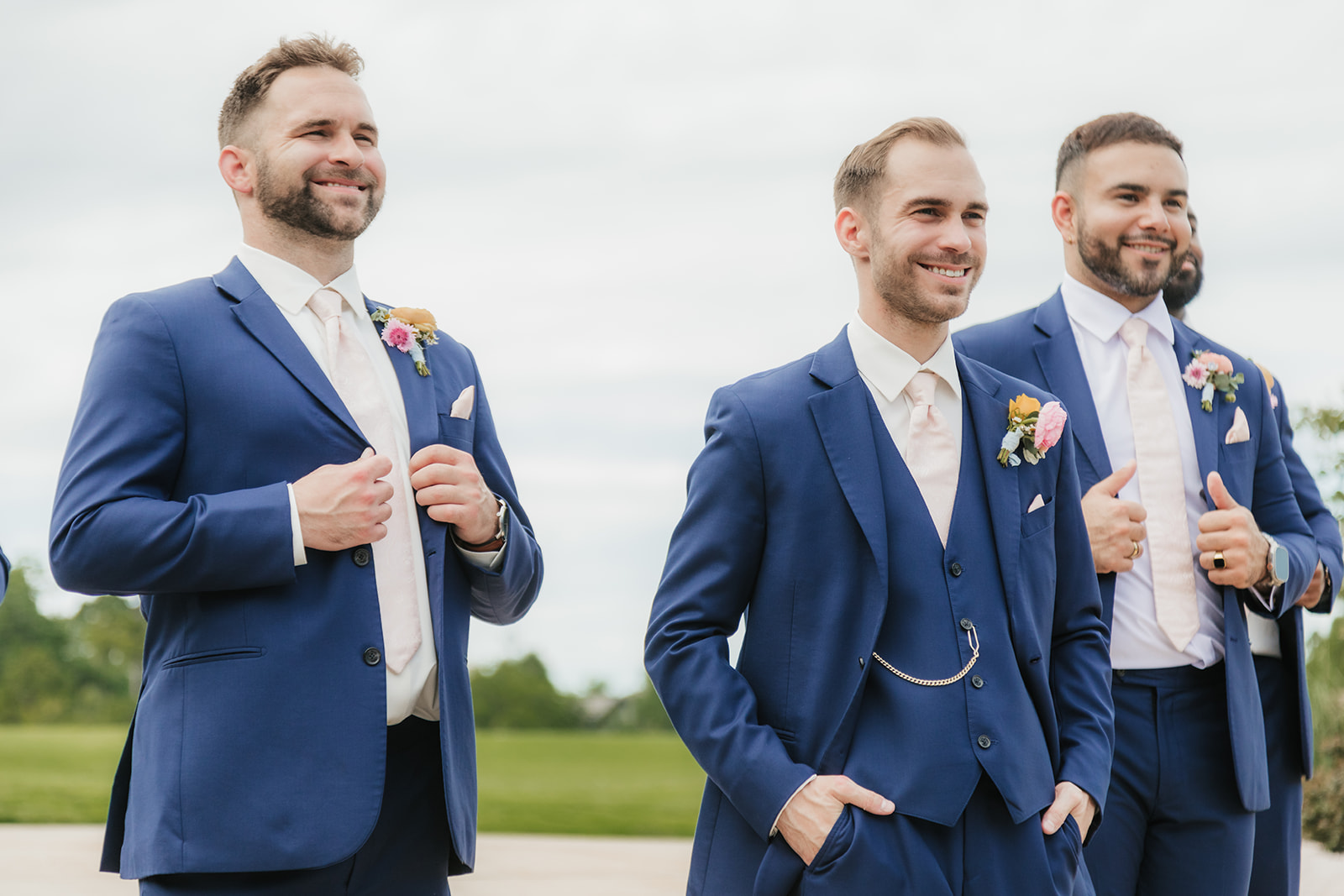 Three men in blue suits with boutonnieres stand outdoors, smiling and posing for a photo on a cloudy day for a Wedding at St. Thomas More University Parish