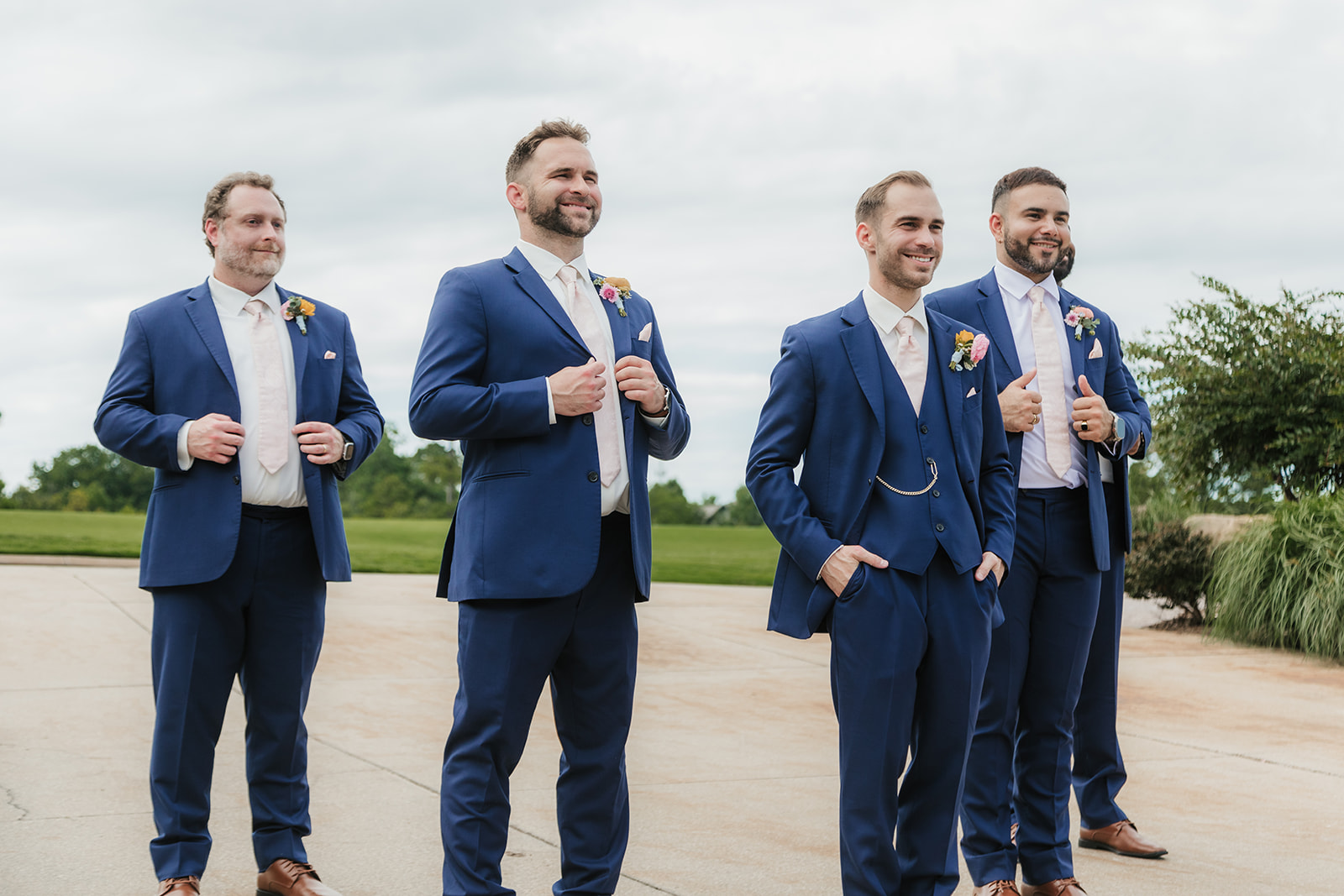 Three men in blue suits with boutonnieres stand outdoors, smiling and posing for a photo on a cloudy day for a Wedding at St. Thomas More University Parish