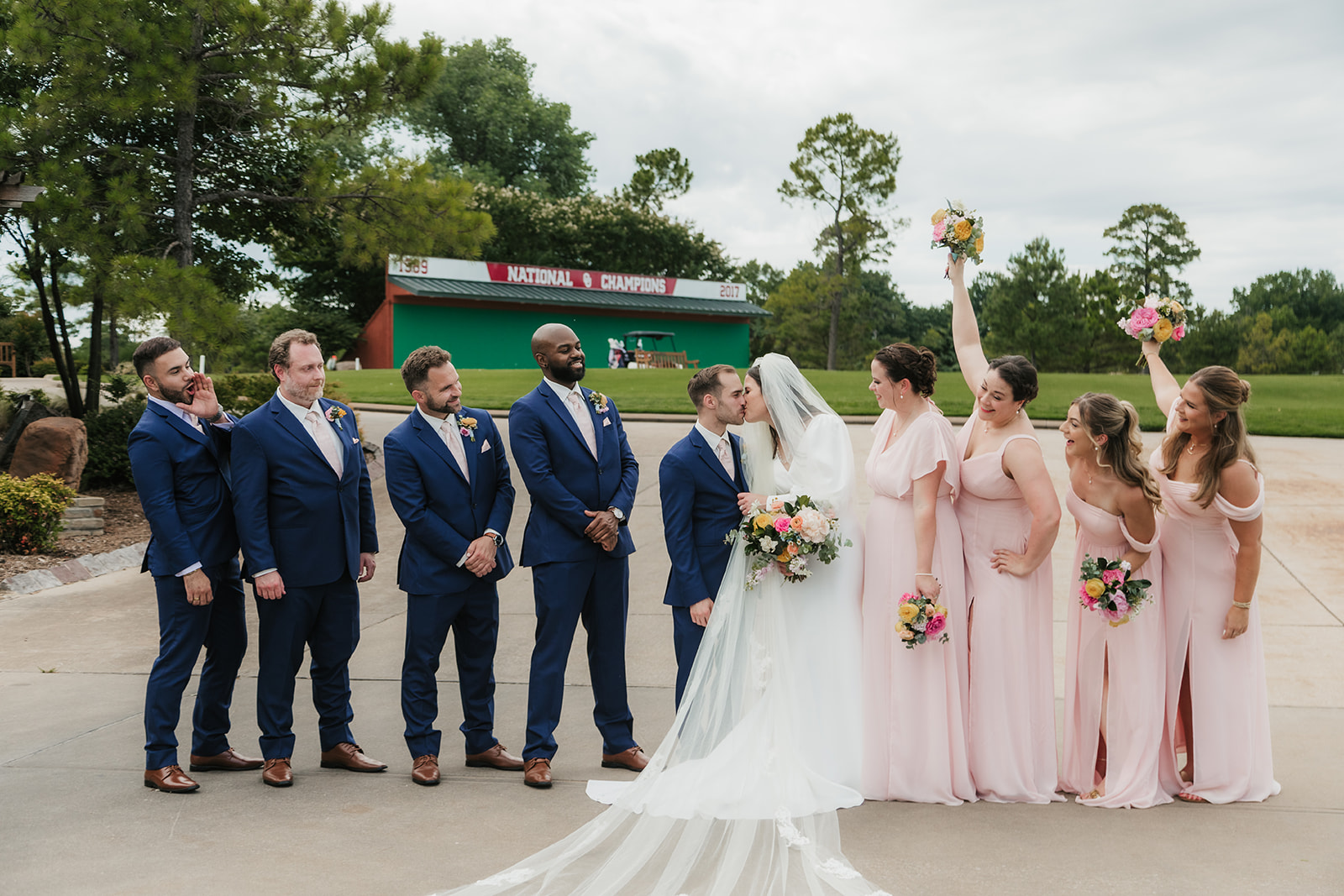 A bride and groom stand with four groomsmen in blue suits and five bridesmaids in light pink dresses, holding bouquets, outdoors on a paved area for a Wedding at St. Thomas More University Parish