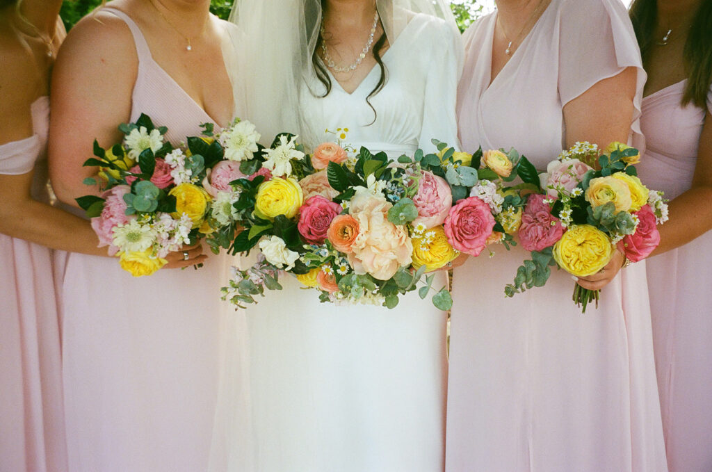 A bride and bridesmaids in light pink dresses hold colorful flower bouquets, featuring yellow, pink, and white blooms.