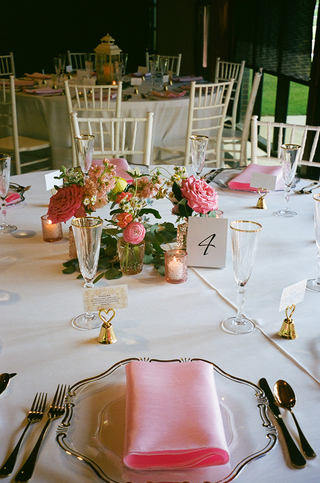 A round table set for a formal event with pink napkins, glassware, floral centerpiece, and a table number 4 card. White chairs and another decorated table are visible in the background.