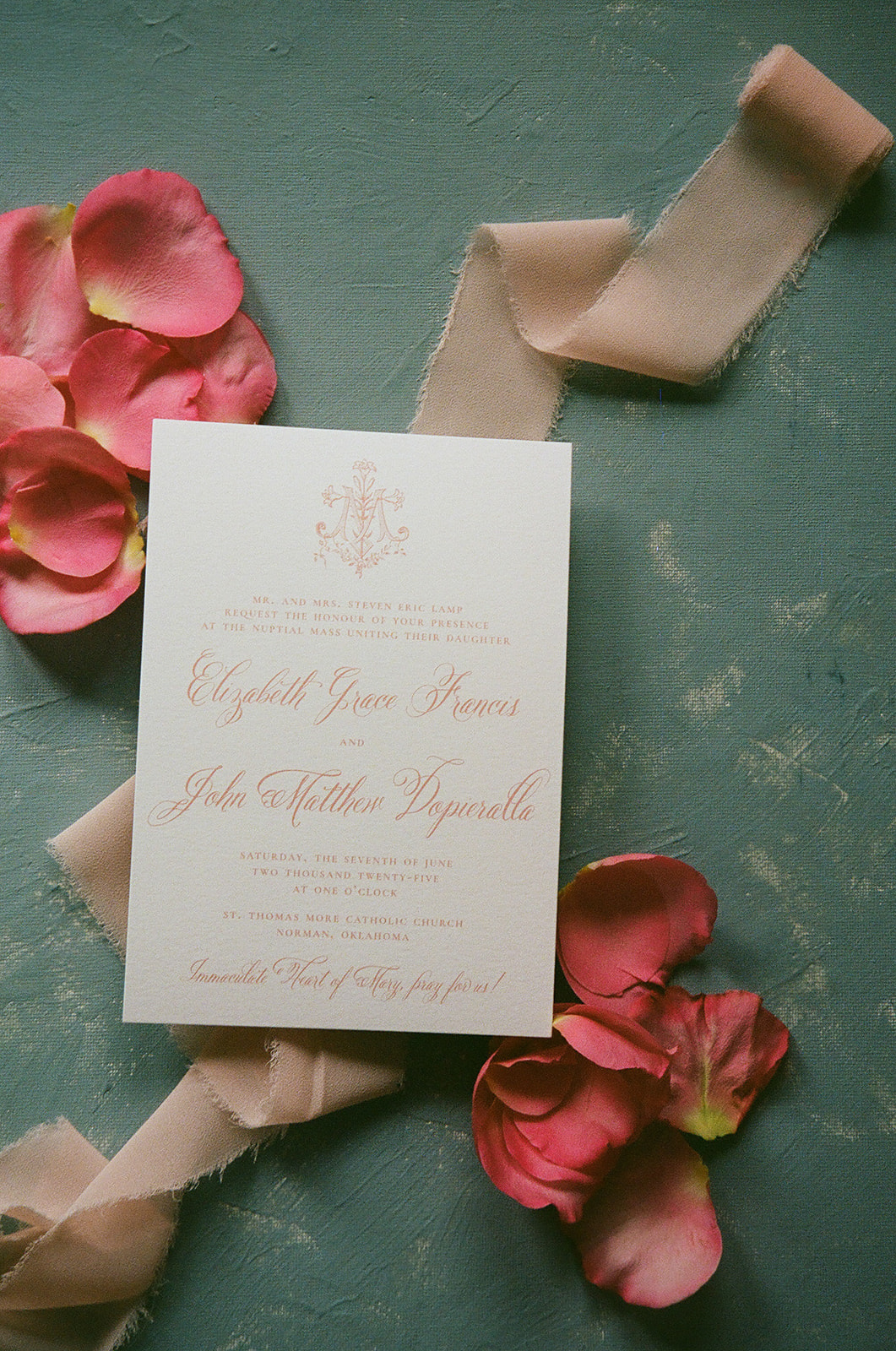 A wedding invitation card with pink script text lies on a blue surface, surrounded by pink rose petals and a beige ribbon.