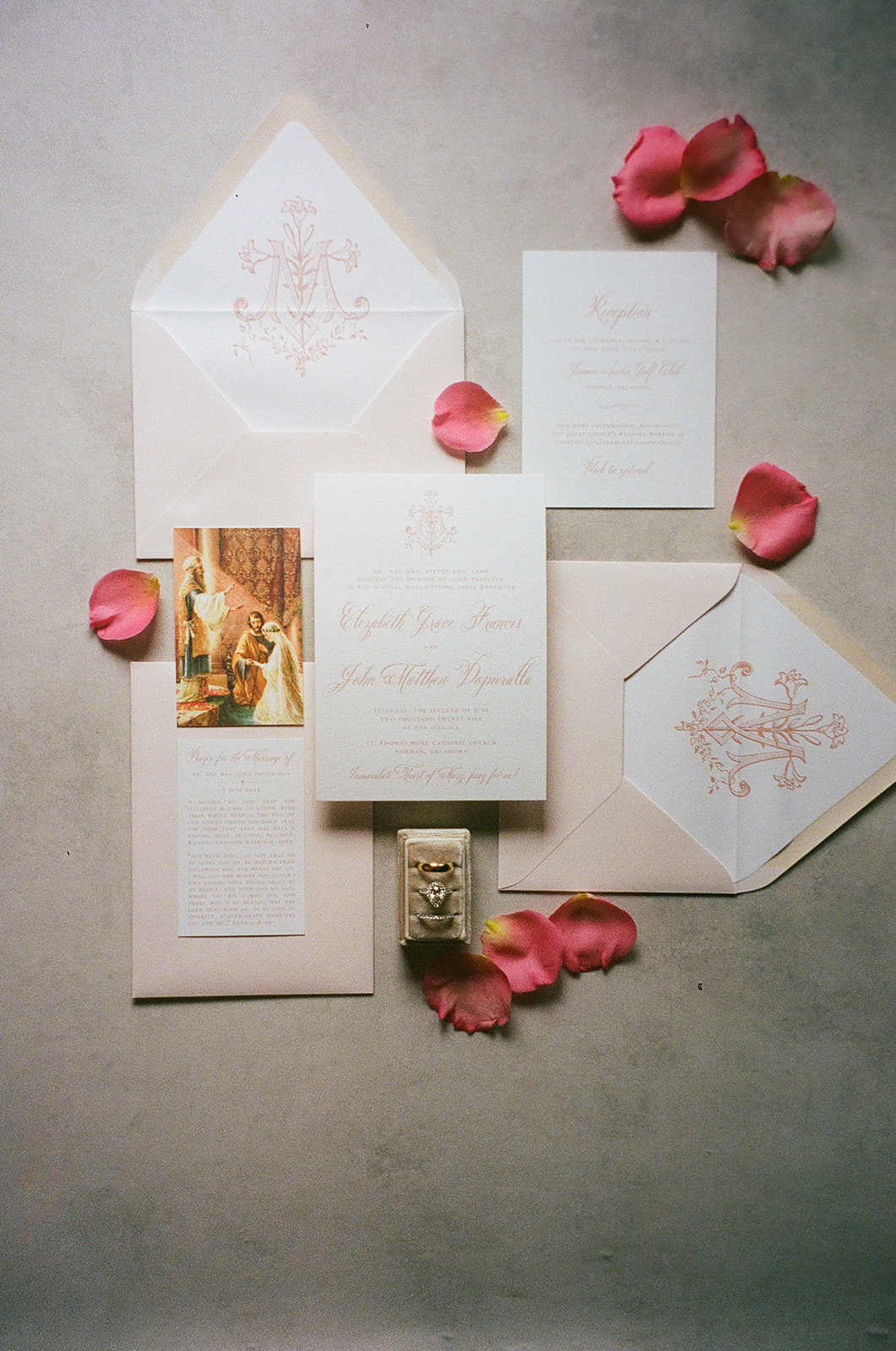 A wedding invitation card with pink script text lies on a blue surface, surrounded by pink rose petals and a beige ribbon for a wedding at