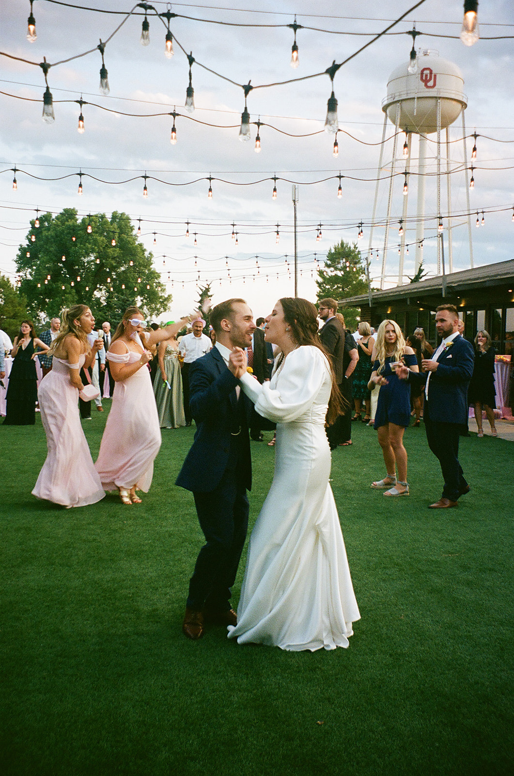 A bride and groom dance outside on green turf under string lights, surrounded by wedding guests, with a water tower visible in the background.