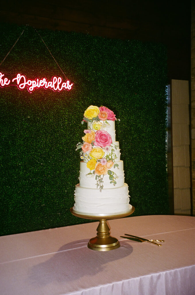A three-tier white cake decorated with colorful flowers sits on a gold cake stand atop a pink tablecloth, with a cake knife nearby and a neon sign in the background.