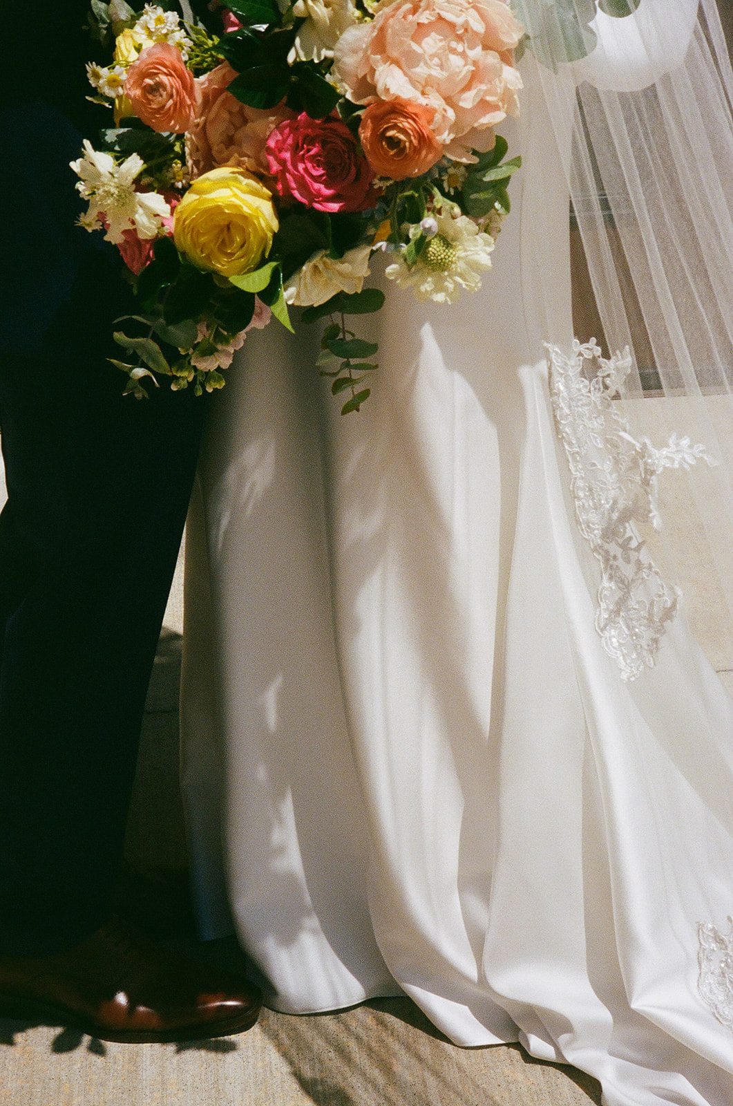 A bride and groom embrace, with the bride holding a bouquet of colorful flowers, including roses, while wearing a white wedding dress and veil.
