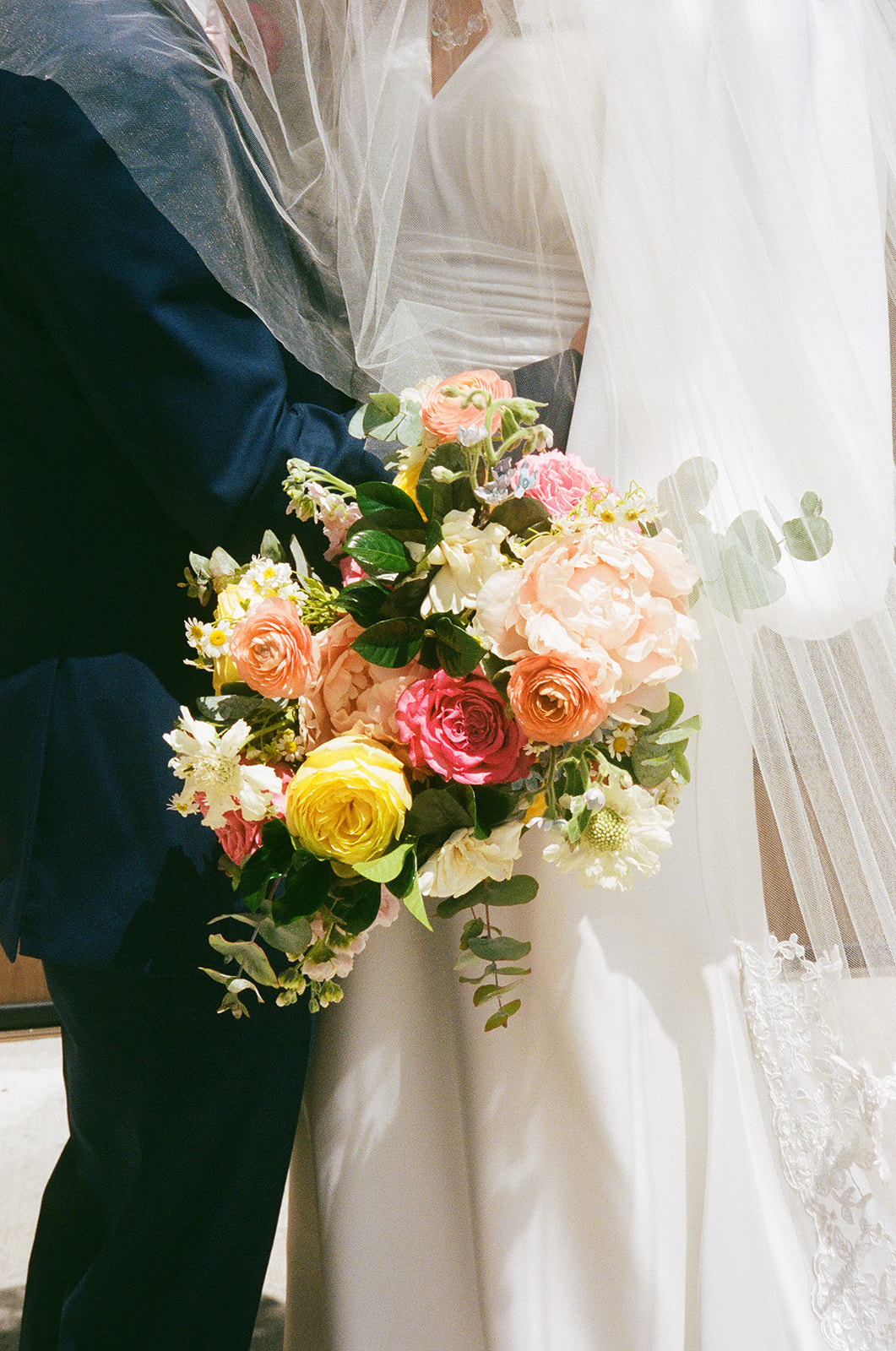 A bride and groom embrace, with the bride holding a bouquet of colorful flowers, including roses, while wearing a white wedding dress and veil.