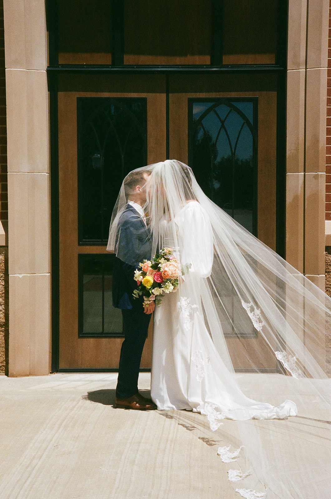 A bride in a white dress and veil stands beside a groom in a blue suit, both smiling and holding a colorful bouquet in front of wooden doors with glass panels for a Wedding at St. Thomas More University Parish