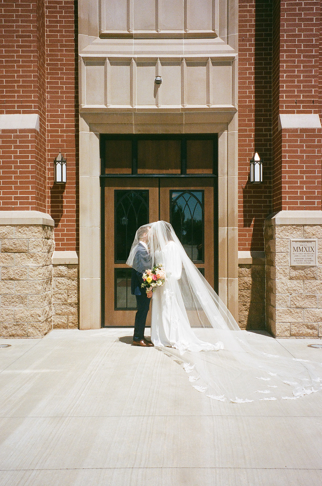 A bride and groom stand in front of a building entrance, sharing a kiss under the bride’s veil outside of St. Thomas More University Parish