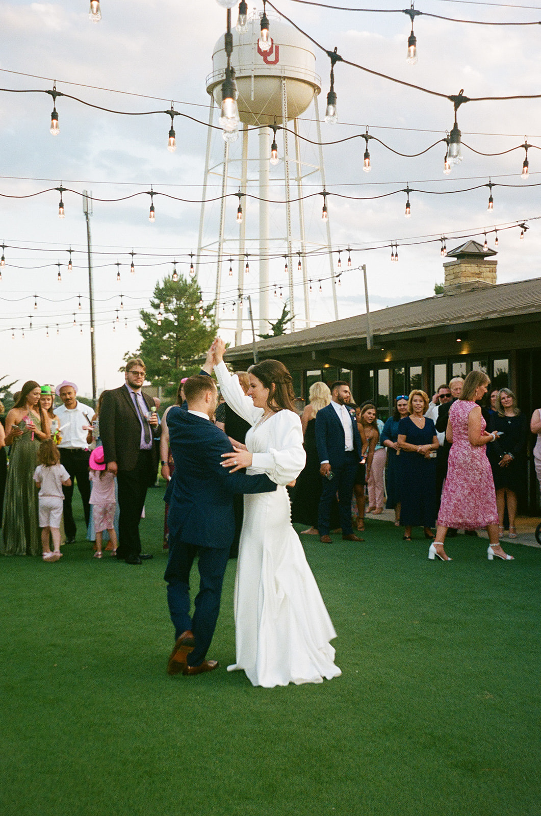 A bride and groom dance outside on green turf under string lights, surrounded by wedding guests, with a water tower visible in the background.