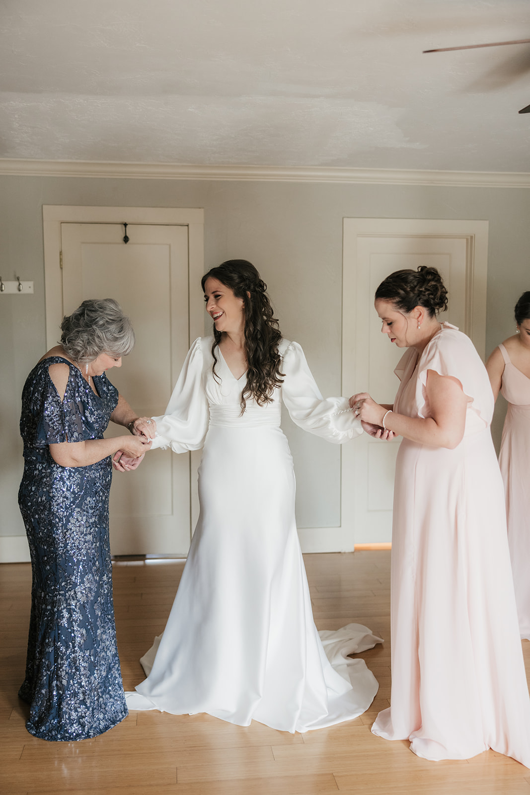 A bride in a white dress stands indoors, holding hands with two women in formal dresses, preparing for a wedding at St. Thomas More University Parish