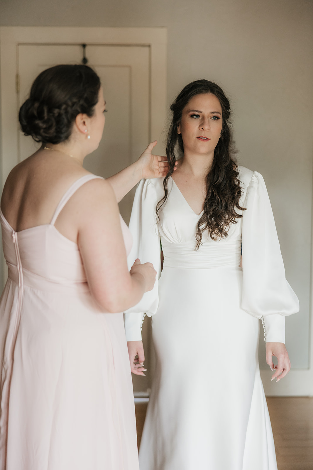 A bride in a white dress stands indoors, holding hands with two women in formal dresses, preparing for a wedding at St. Thomas More University Parish
