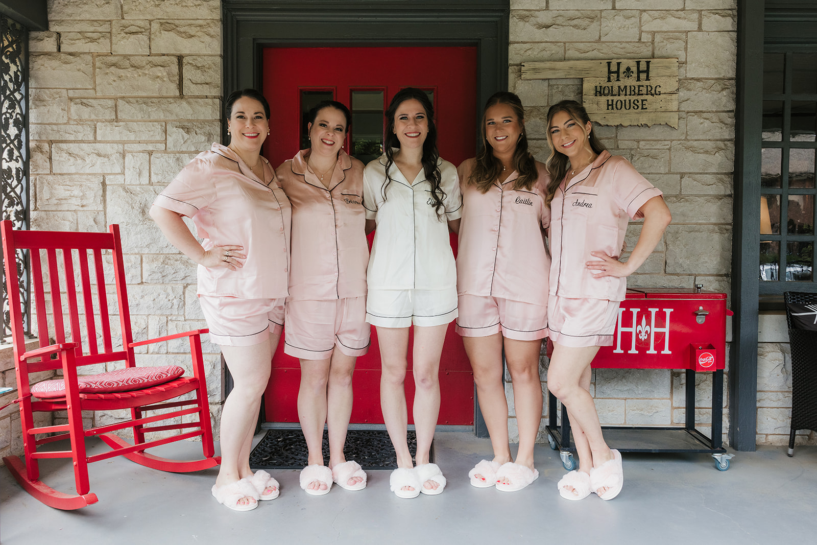 Five women stand side by side in matching pajamas and slippers in front of a red door, with a red rocking chair and a cooler to the right.
