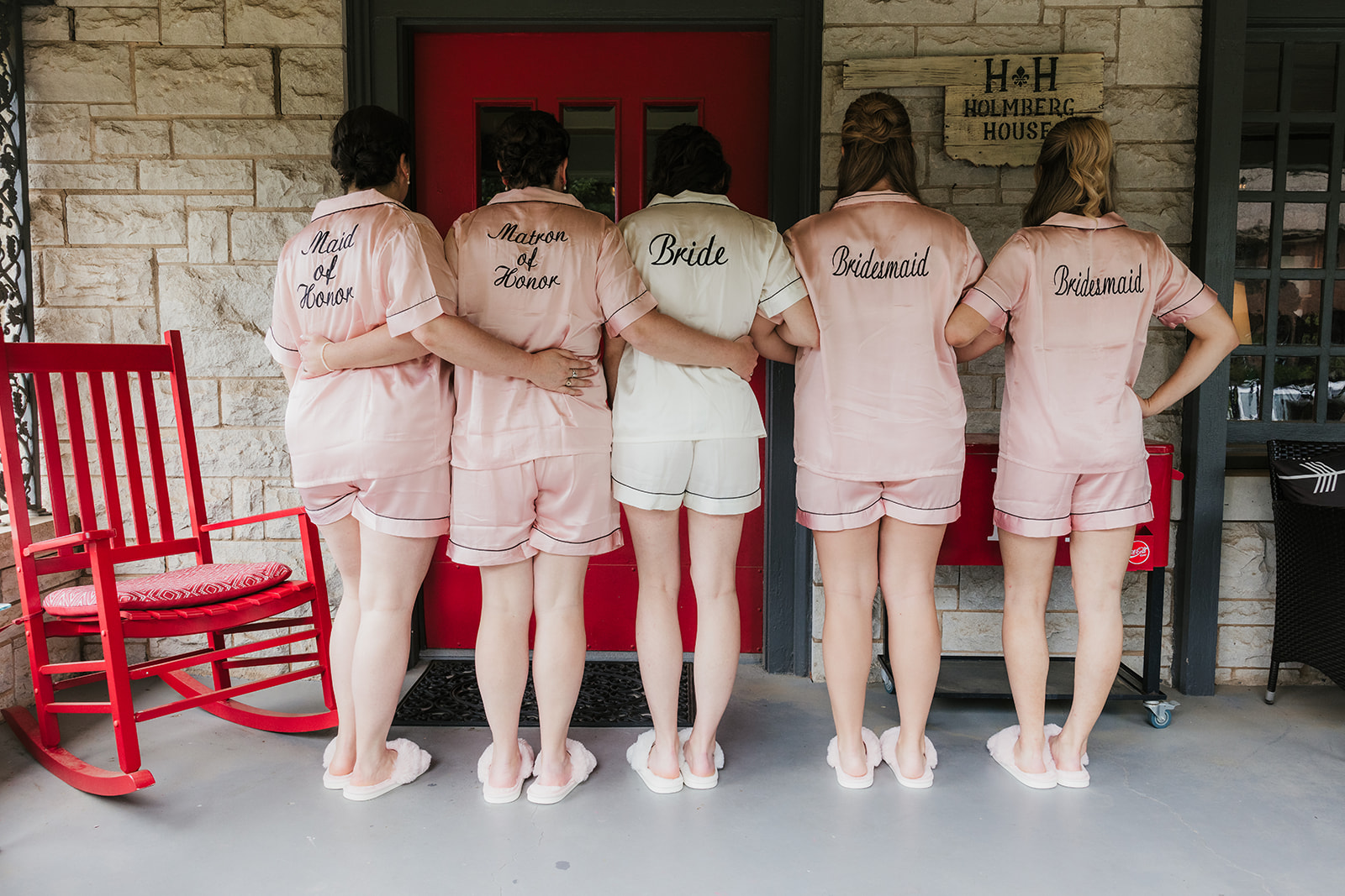 Five women stand side by side in matching pajamas and slippers in front of a red door, with a red rocking chair and a cooler to the right.