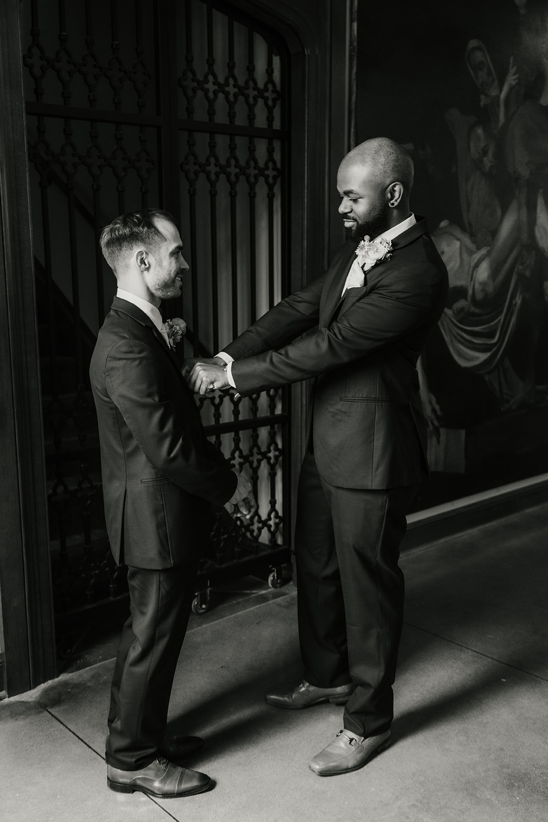 Two men in suits stand facing each other indoors, one adjusting the other's cufflinks. The background includes a decorative gate and a large mural.
