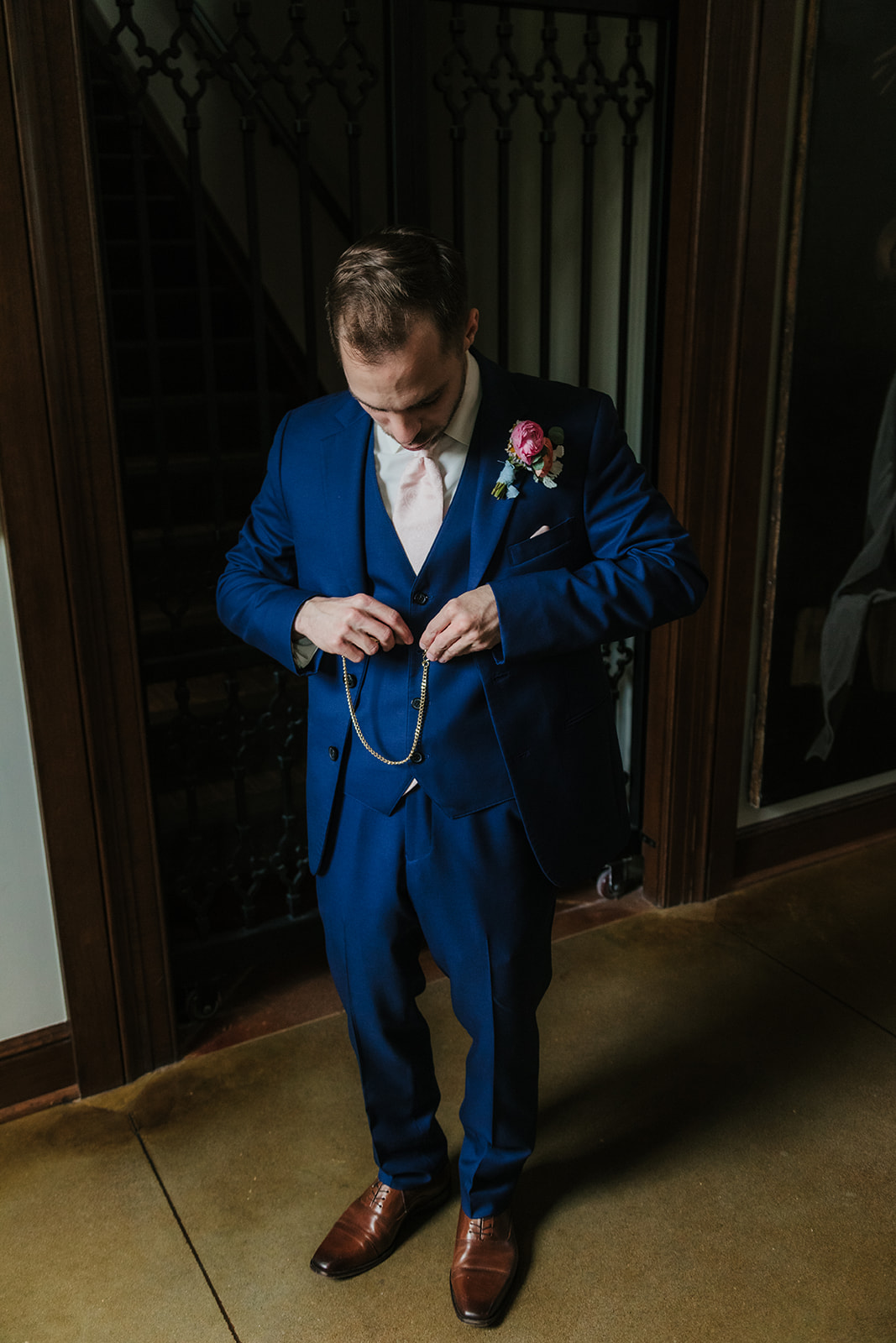 A man in a blue suit stands indoors, looking down while fastening a pocket watch chain.