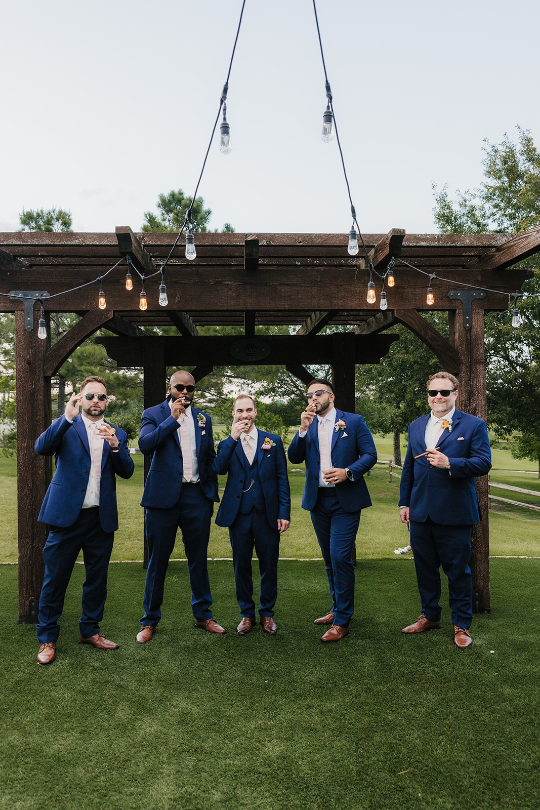 Five men in blue suits pose under a wooden pergola outdoors, some adjusting ties or sunglasses, on a lawn with trees in the background.