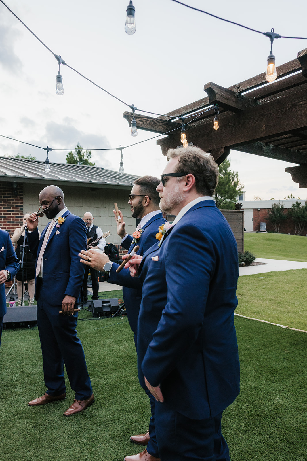 Four men in matching blue suits stand on grass under string lights at an outdoor event, with a pergola and buildings in the background.