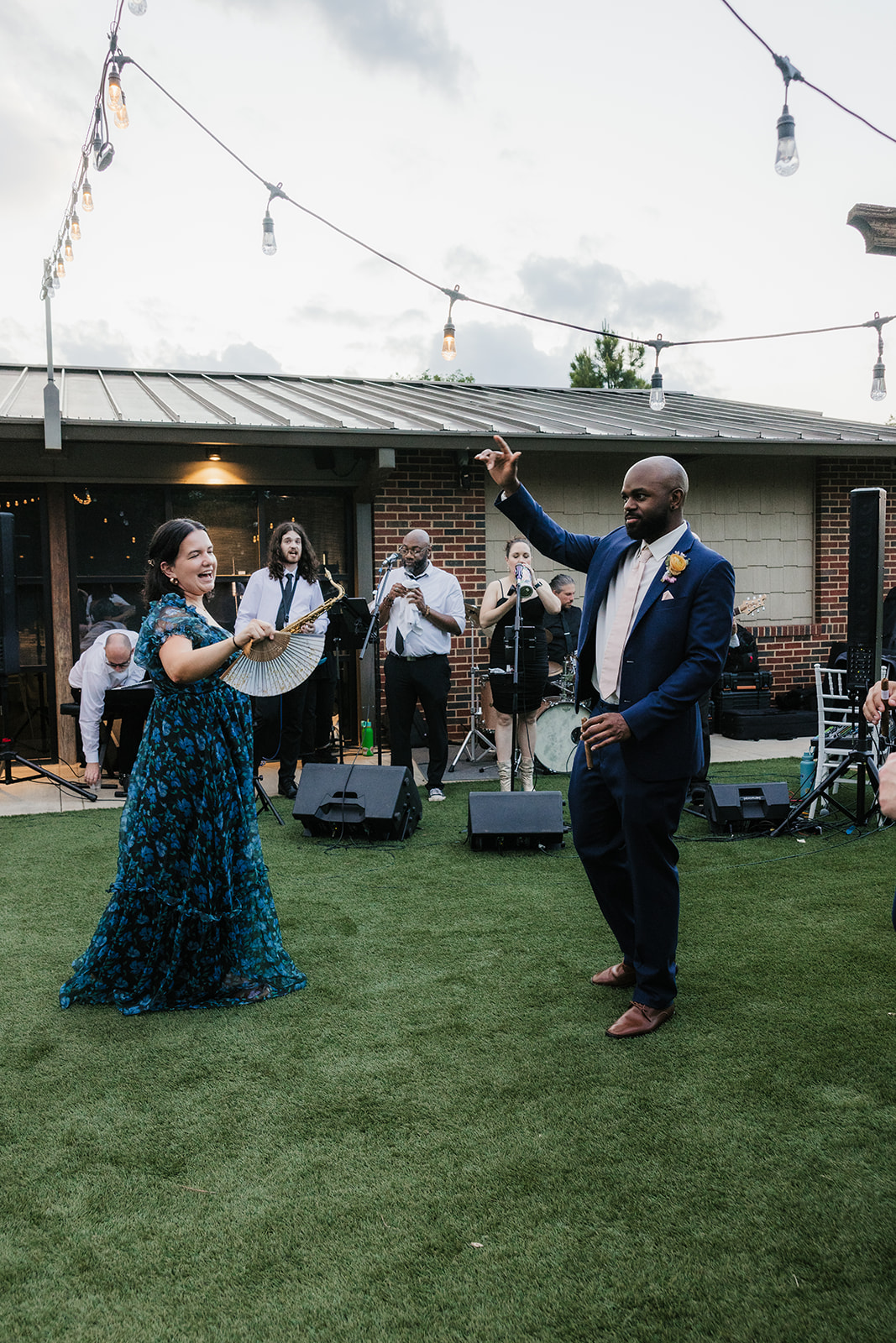 A woman and a man dance together on a lawn during an outdoor event, with musicians and guests watching in the background.