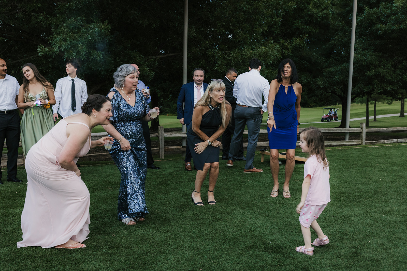 A group of adults in formal attire playfully interact with a young girl on a grassy lawn at an outdoor gathering.