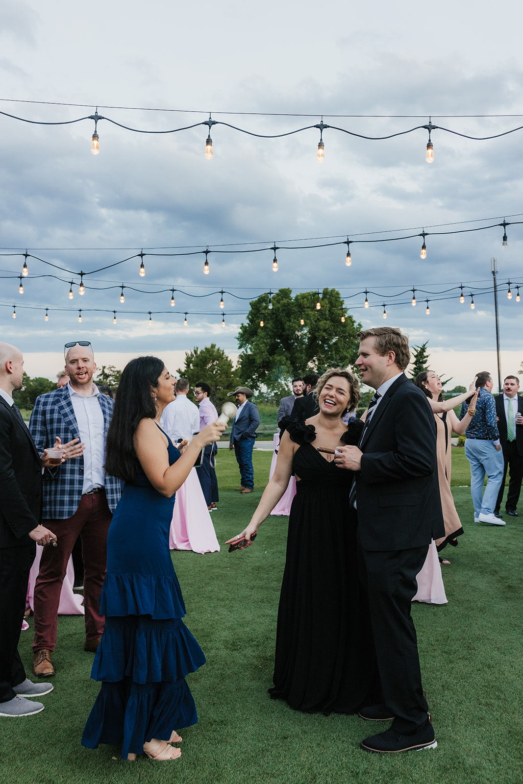 A group of adults in formal attire playfully interact with a young girl on a grassy lawn at an outdoor gathering.