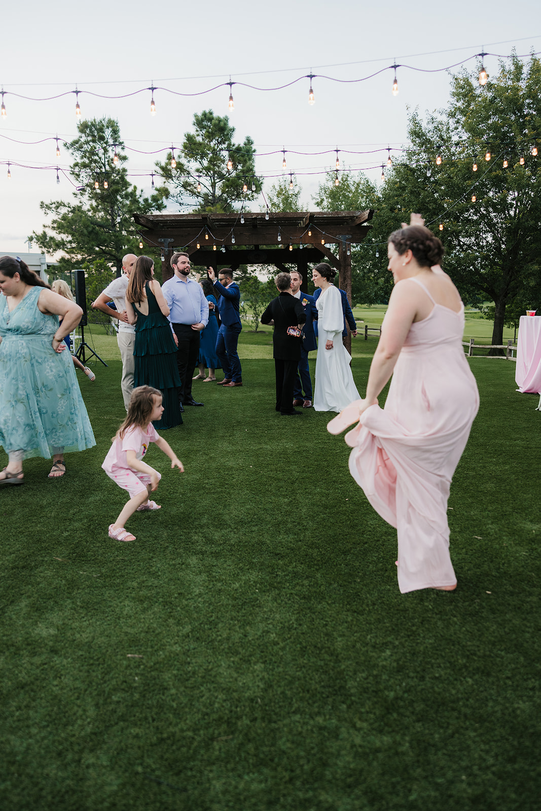 A group of adults in formal attire playfully interact with a young girl on a grassy lawn at an outdoor gathering.
