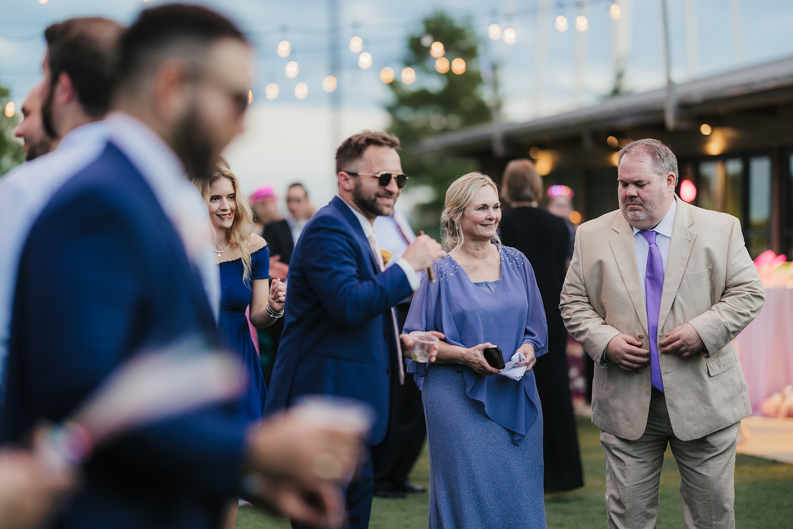 A group of adults in formal attire playfully interact with a young girl on a grassy lawn at an outdoor gathering.